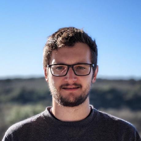 A man with short brown hair, black glasses, and a beard standing outdoors against a blurred landscape and blue sky.
