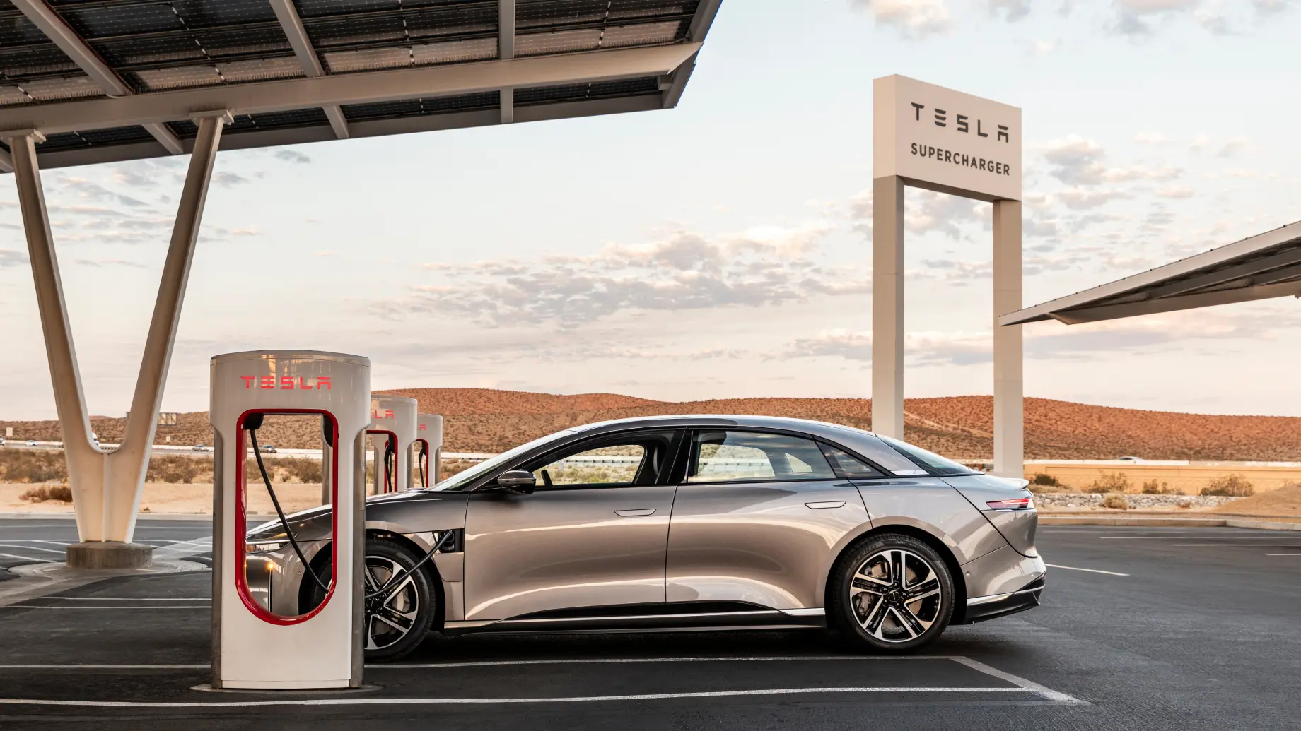 Lucid Air charging at a Tesla Supercharger station with desert hills in the background under cloudy sky.