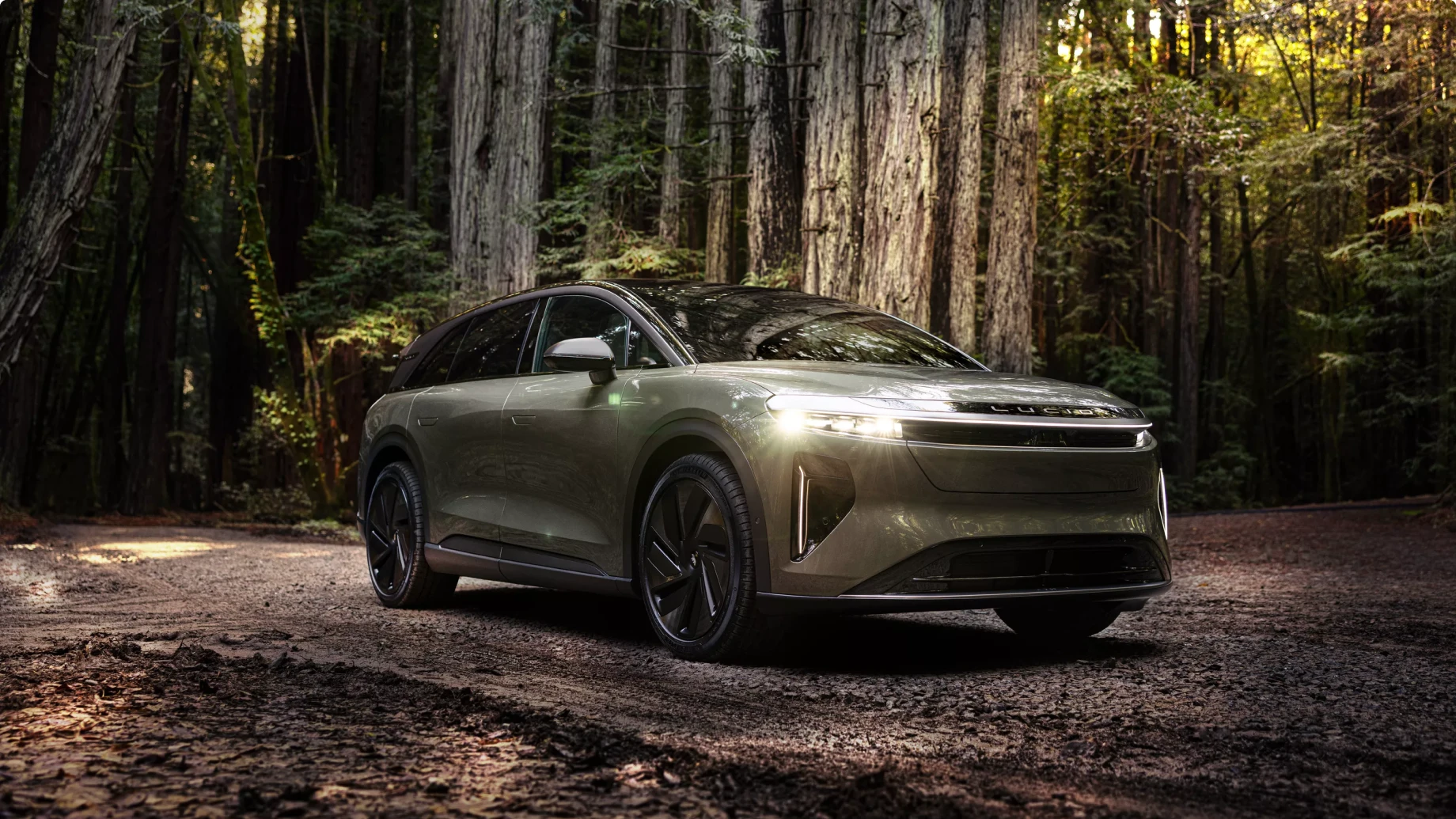 Lucid Gravity with headlights on, parked on forest path surrounded by tall redwood trees in dappled light.
