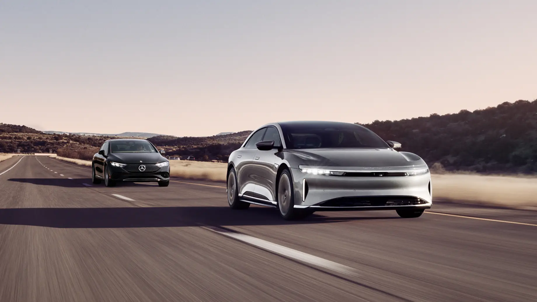A Lucid Air leads a dark Mercedes on a desert highway, with arid hills and a hazy sky in the background.