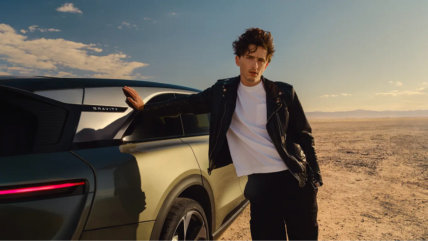 Timothée Chalamet in black leather jacket leaning against sleek modern Lucid Gravity in desert landscape under blue sky