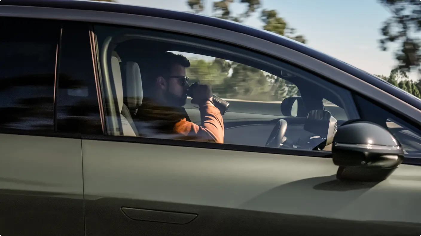 Un homme portant une veste orange boit dans une tasse de voyage tout en étant assis sur le siège passager d’une voiture verte en mouvement.