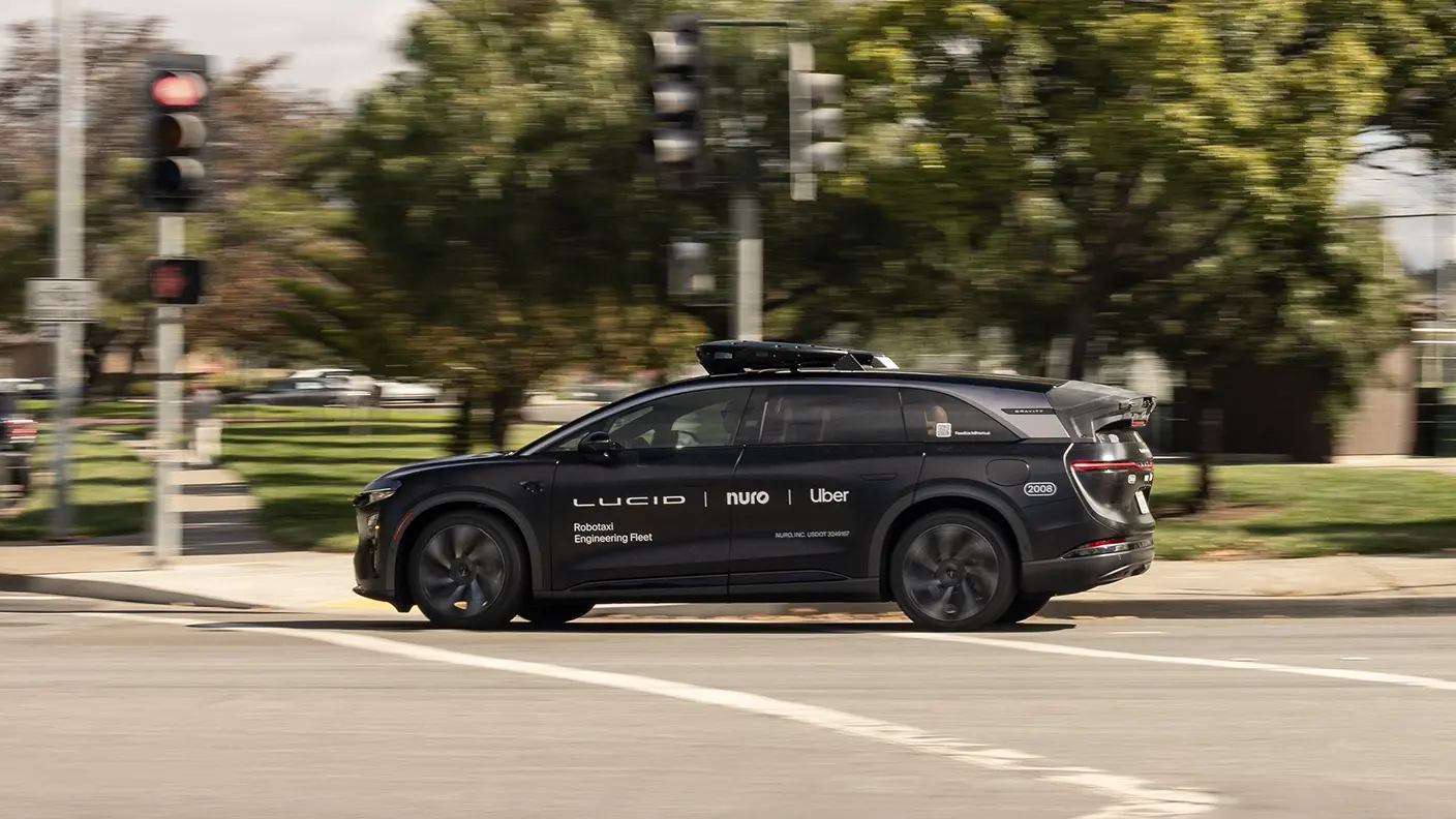 Black Lucid autonomous vehicle with Uber and Nuro branding driving through an intersection with traffic lights and trees.