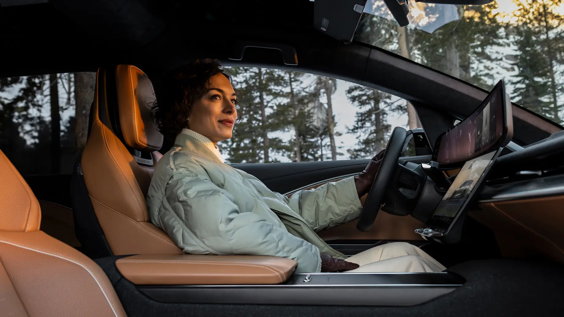 Woman in a light jacket sitting in a modern car with tan leather interior and a large touchscreen, trees visible through the windshield.