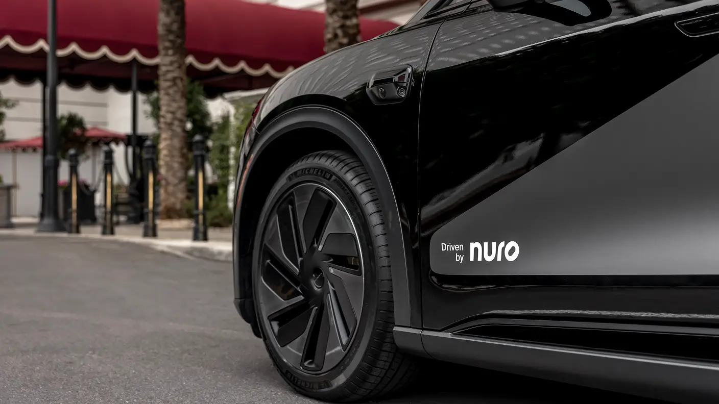 Close-up of a sleek black vehicle with "Driven by nuro" logo on its side, parked near a building with red awnings.