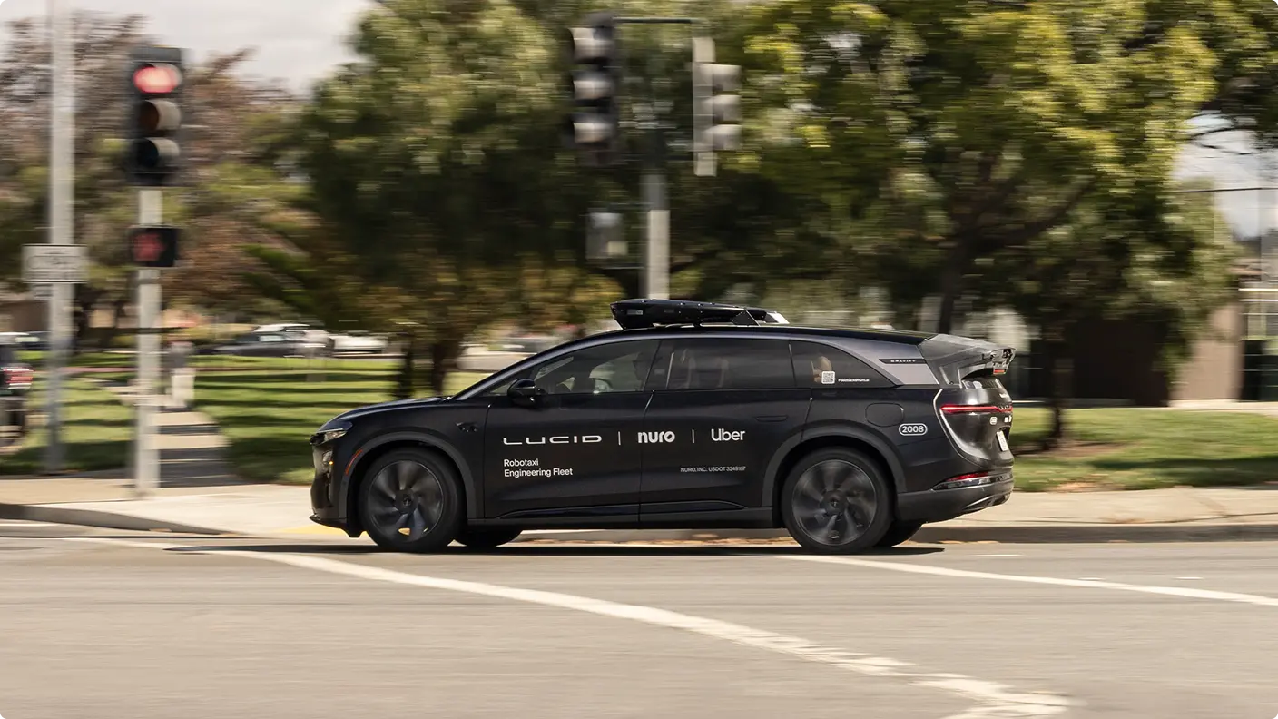 Black Lucid, Nuro, and Uber robotaxi engineering fleet vehicle with rooftop sensors driving through a suburban intersection.