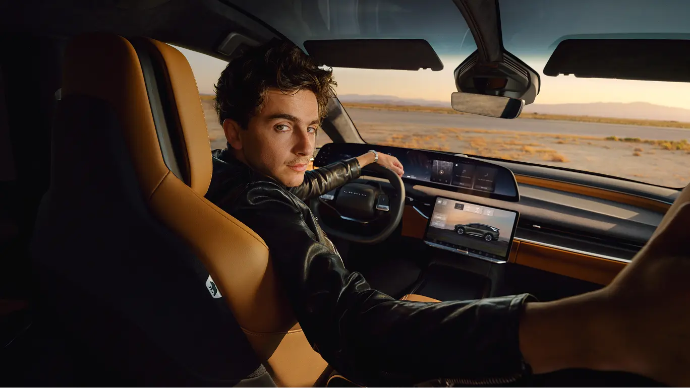 Timothée Chalamet driving a Lucid vehicle with tan leather interior through a desert landscape at sunset, digital dashboard visible.